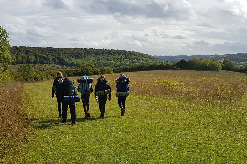 Five pupils walking through a field whilst wearing large backpacks.