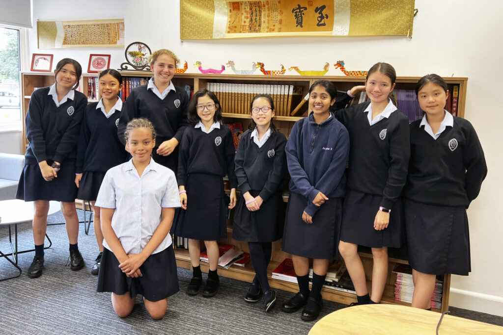 Pupils in front of bookcase. On top of the bookcase are multicoloured paper dragon boats, above is a gold banner with Chinese writing.