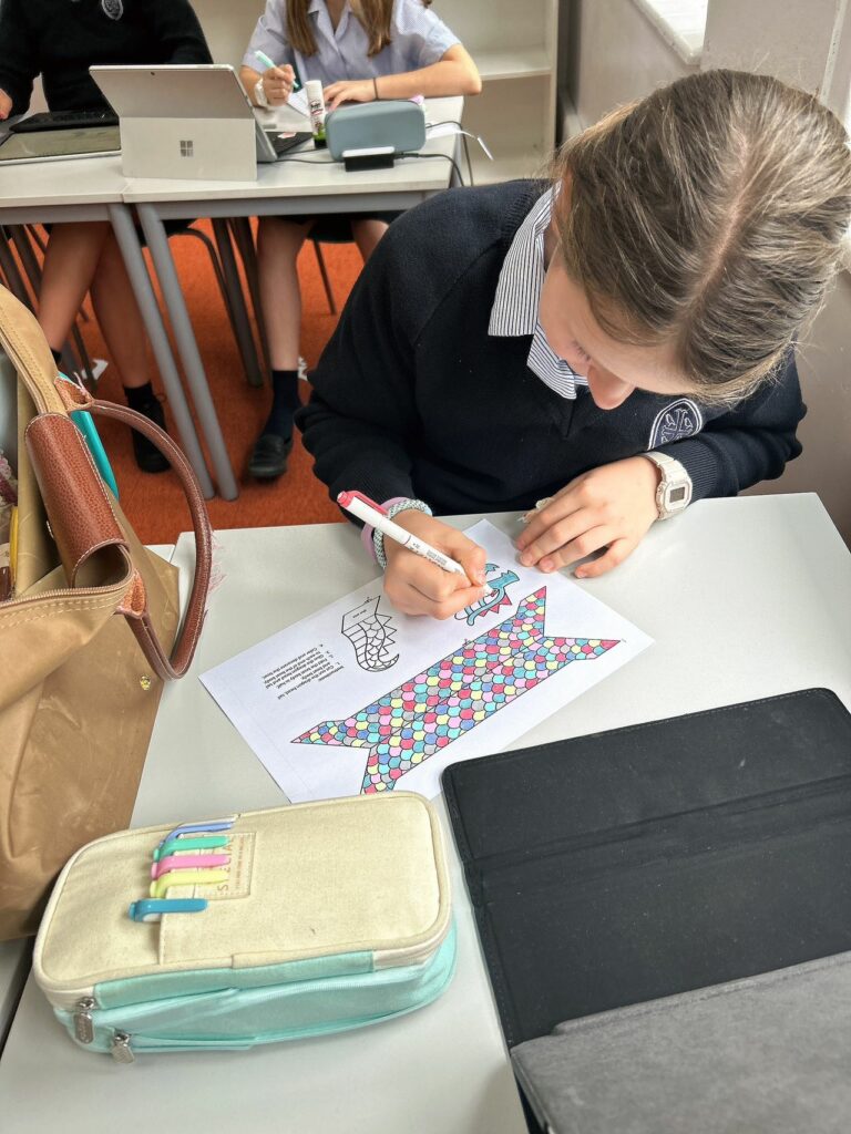 A pupil colouring in her dragon boat. her pencil case and laptop are open in front of her. Her light brown bag is to the right of her.