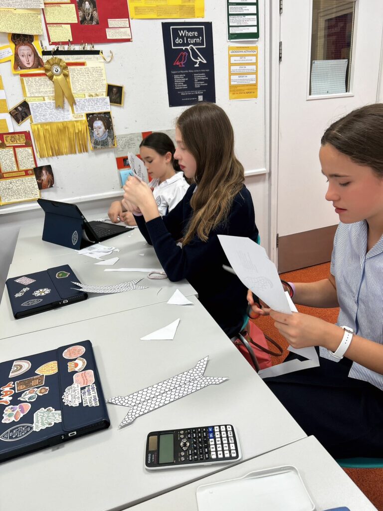 Three pupils sat at a table with a laptop in front of them. They are cutting out elements needed for their dragon boat.