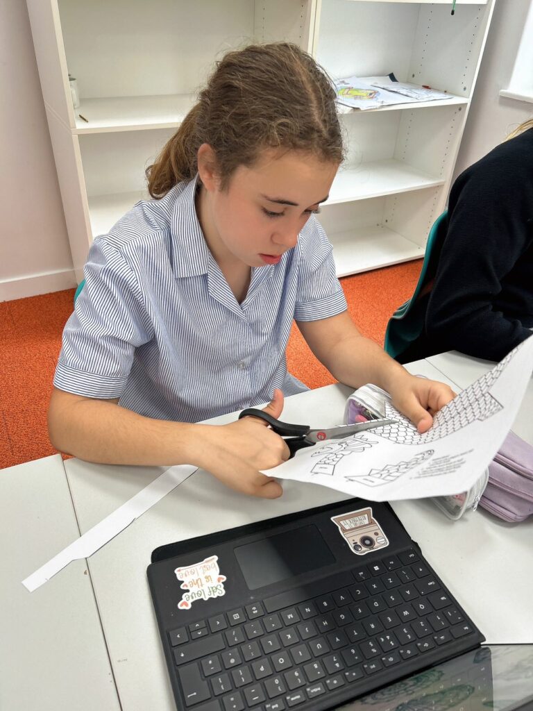 A pupil sat at a table with a laptop in front of her. She is cutting out elements needed for her dragon boat.