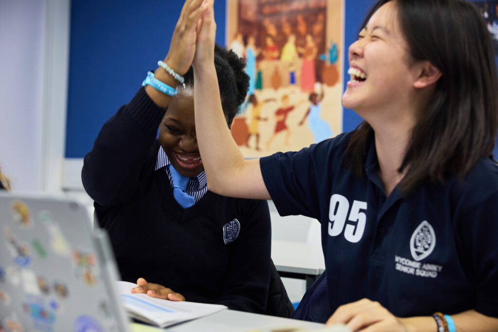 Two pupils high fiving one another whilst sat at their desks with a laptop and textbook.