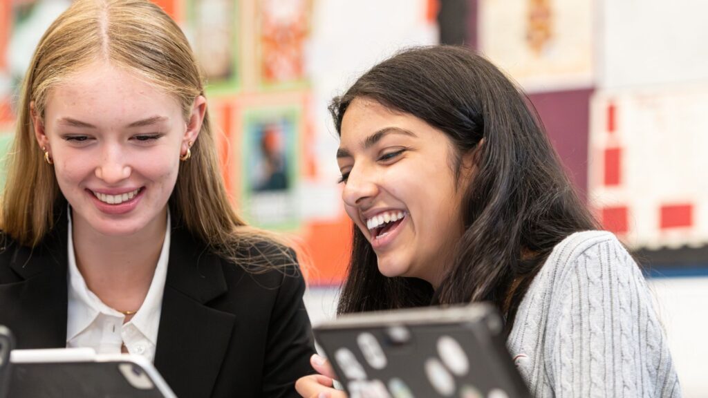 Two pupils smiling and laughing whilst looking at a laptop.