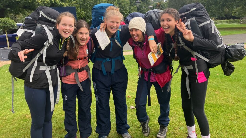 Five pupils with their hand round one another smiling whilst wearing large backpacks.