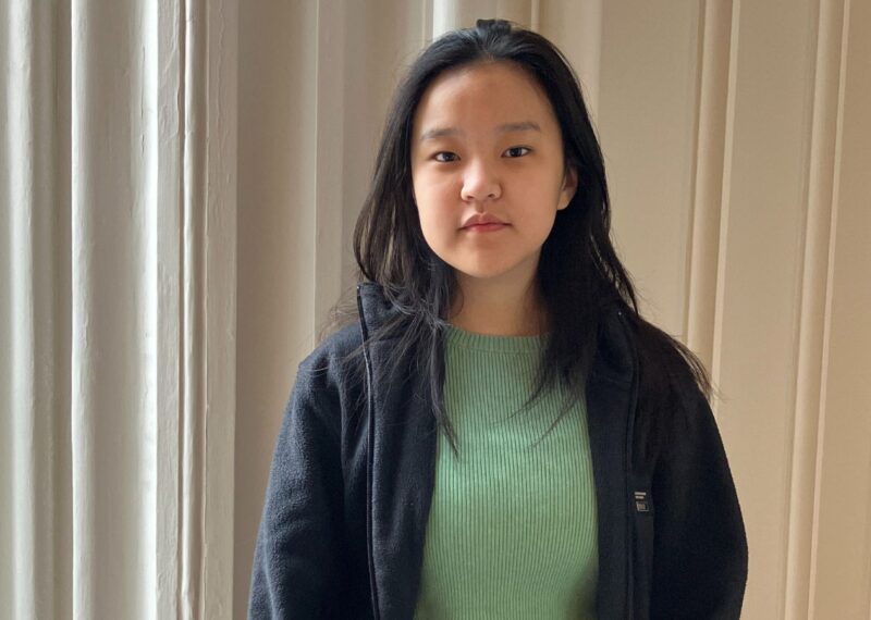 A young woman wearing a green shirt and a black jacket, standing confidently with a neutral expression.