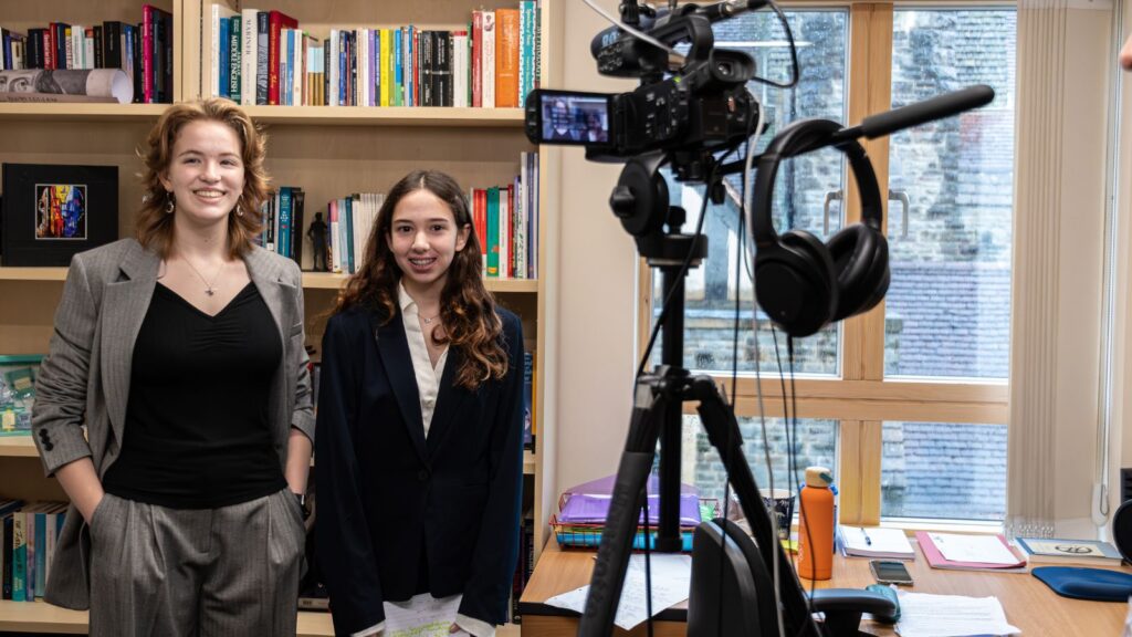 Two Sixth Form pupils dressed in suits, smiling in front of a video camera.