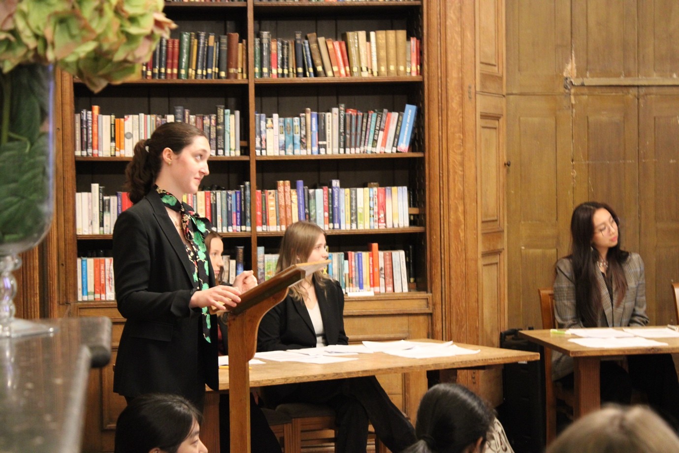 A pupil with raised eyebrows stands at a lectern. Three pupils are sat at desks to her left.
