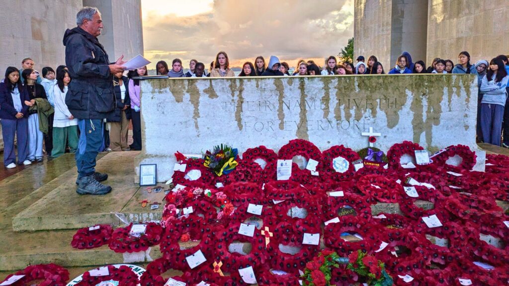 A man stood giving a speech in front of pupils. Lots of poppy wreaths are leant against a war memorial.