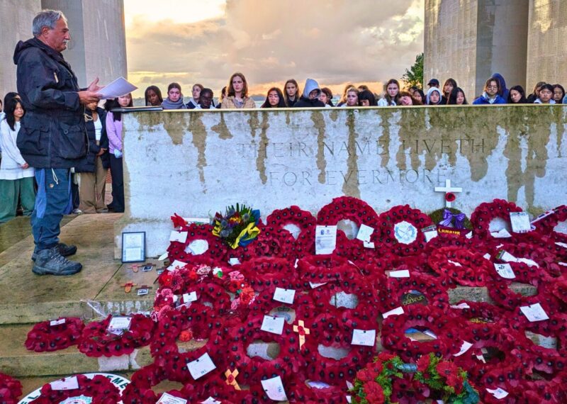An elderly man stood to the side of a WW2 memorial adorned with poppy wreaths. Young women surround the memorial.