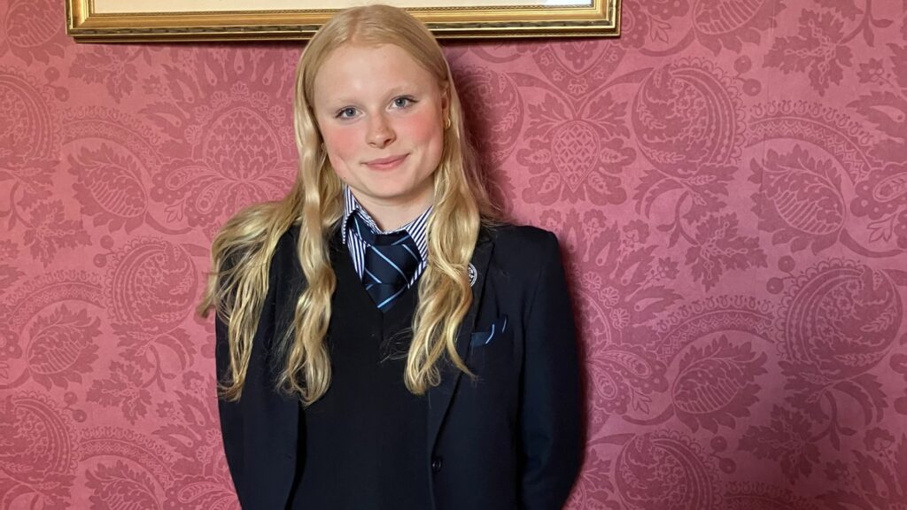 Annabelle smiling in her School uniform, stood in front of a red patterned wall.