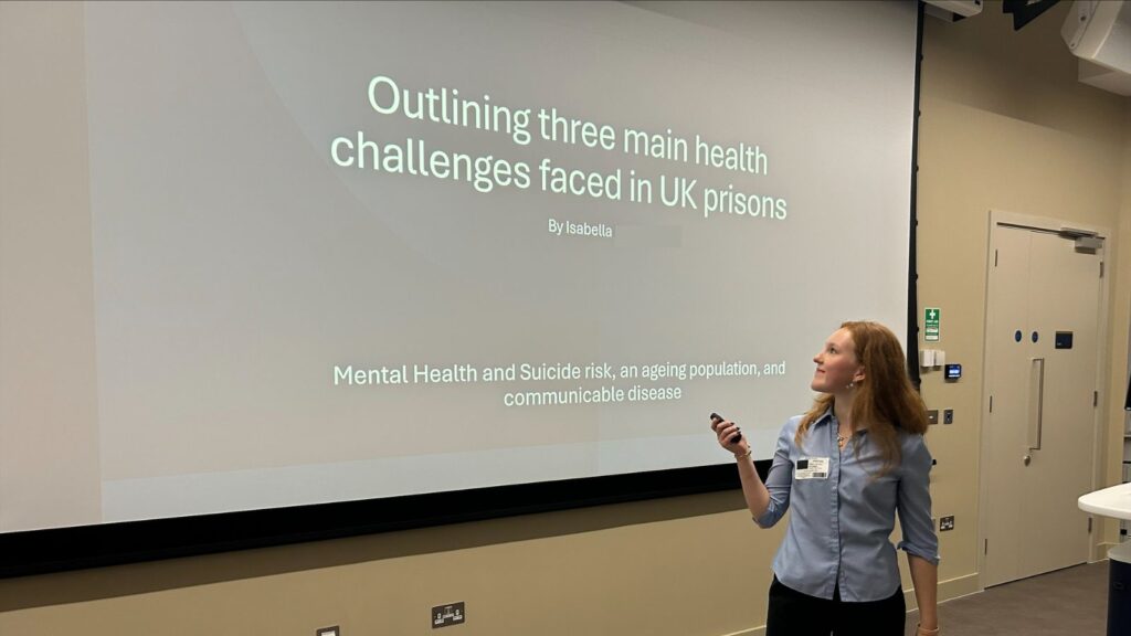 Izzy changing the slide on her presentation titled 'Outlining three main health challenges faced in UK prisons'.
