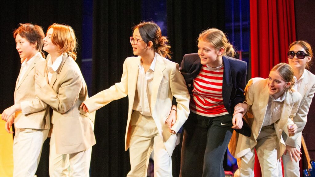 Six Wycombe Abbey pupils, on stage in costumes, looking to the right whilst smiling.