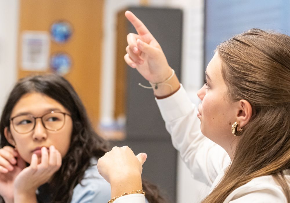 A girl wearing glasses is look at the pupil next to her who has her hand up.