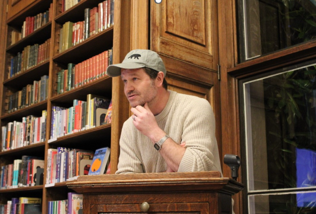 A male teacher wearing a grey cap and a silver watch stands behind a lectern, in front if a book shelf. His left hand is scratching his chin as he thinks.
