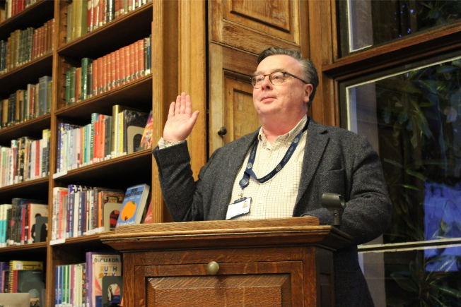 Mr Reid, wearing his lanyard and glasses, stands behind the lectern, in front of the bookcase, with his right hand raised.