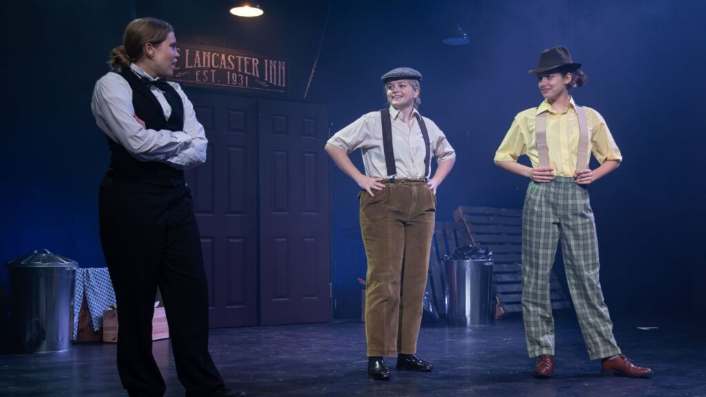 Three girls in male costumes. One in a black suit has their arms crossed and the two wearing suspenders have their hands on their hips