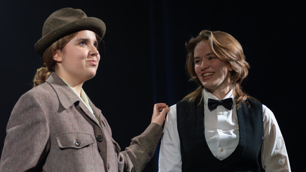 Two girls in male costumes, The girl on the left has her hand on the girl on the right's shoulder.