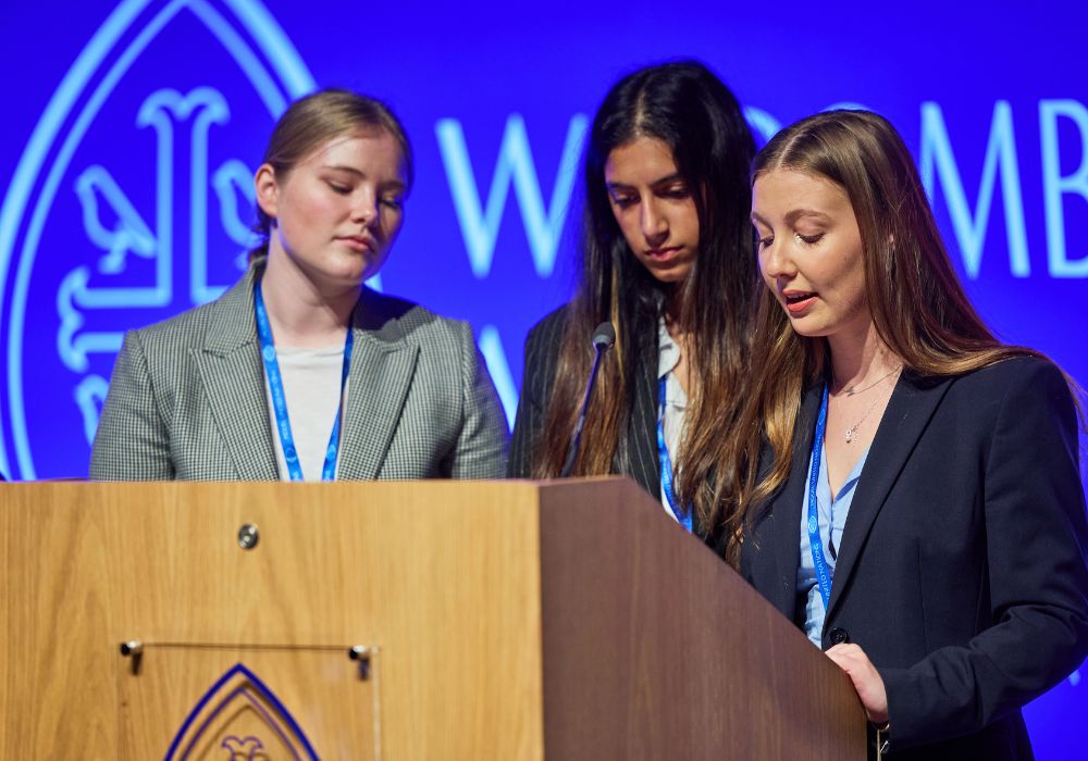 Three girls stood behind a Wycombe Abbey Lectern, in front of the Wycombe Abbey logo.