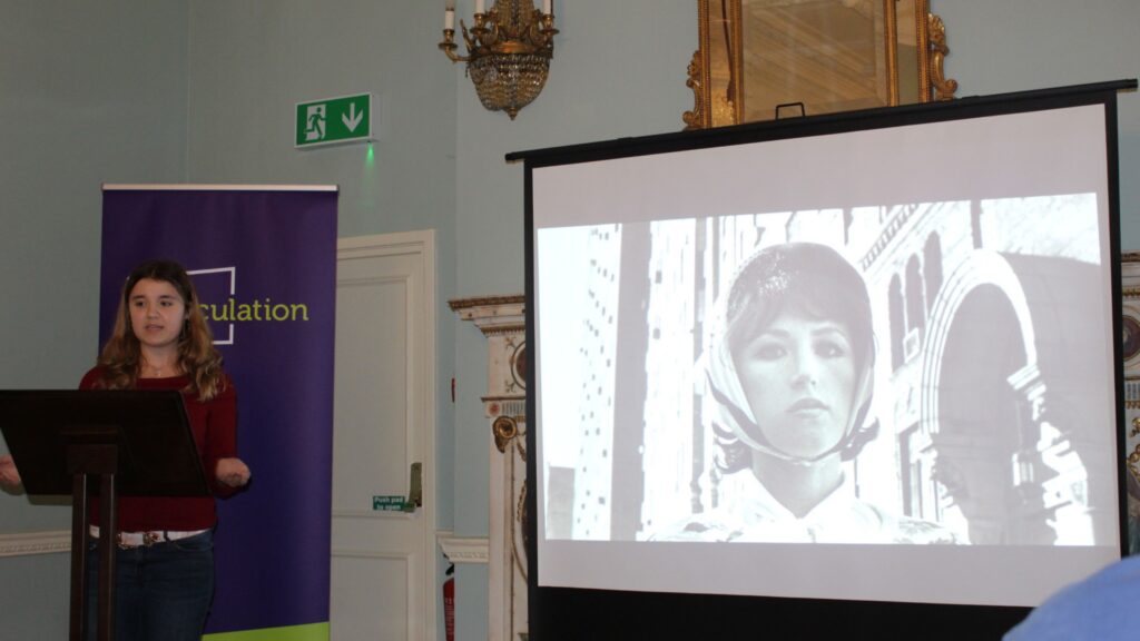 Sasha stood behind a lectern whilst presenting. A black and white picture of a woman wearing a headscarf is on the screen behind her.