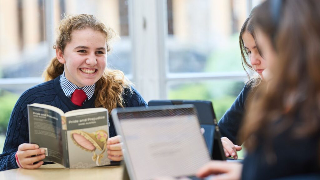 A girl reading a book is smiling across the table at her friend who is on a laptop.