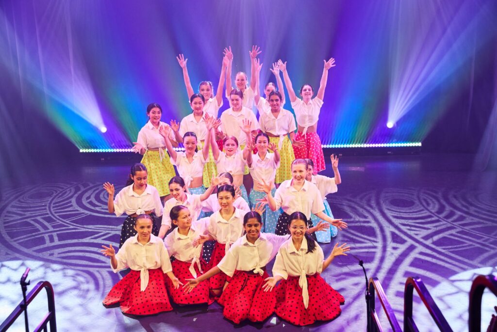 Pupils on stage wearing white shirts with different colour polka dot skirts.