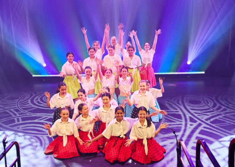 Pupils on stage wearing white shirts with different colour polka dot skirts.