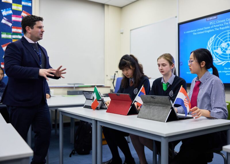 A teacher speaking to three pupils. The pupils each have a country flag representing the country they are debating on behalf of.