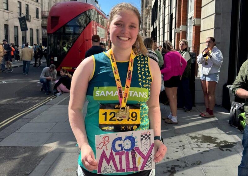 Milly wearing a running top with 'Samaritans' on the front with a 'finisher' medal around her neck. She is holding a handmade poster that reads 'Go Milly'.