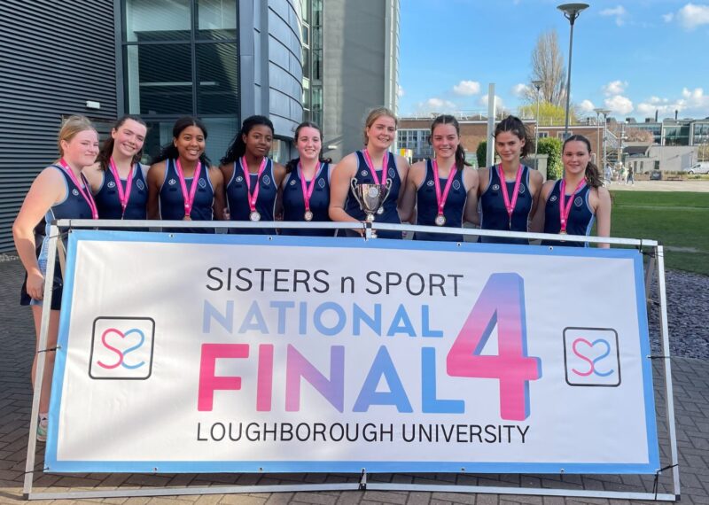 The Wycombe Abbey U19 netball team stood wearing their 1st place medals behind a Sisters n Sport sign.