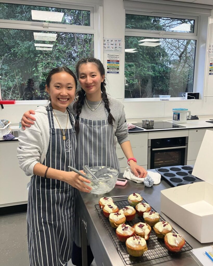 Two girls with their arms round one another, stood behind the cupcakes they have made. The girl on the left on holding a mixing bowl