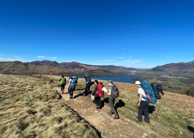 Pupils wearing backpacks, walking amongst hills and lakes.