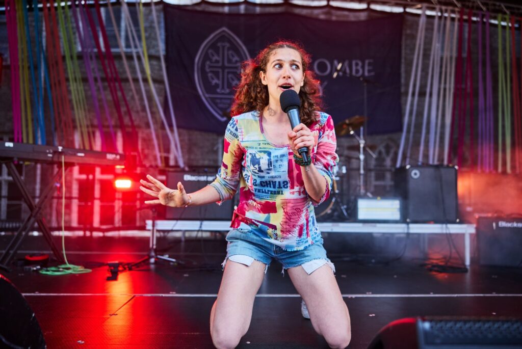 A pupil wearing a multi-coloured top and blue denim shorts is kneeling whilst singing on stage.