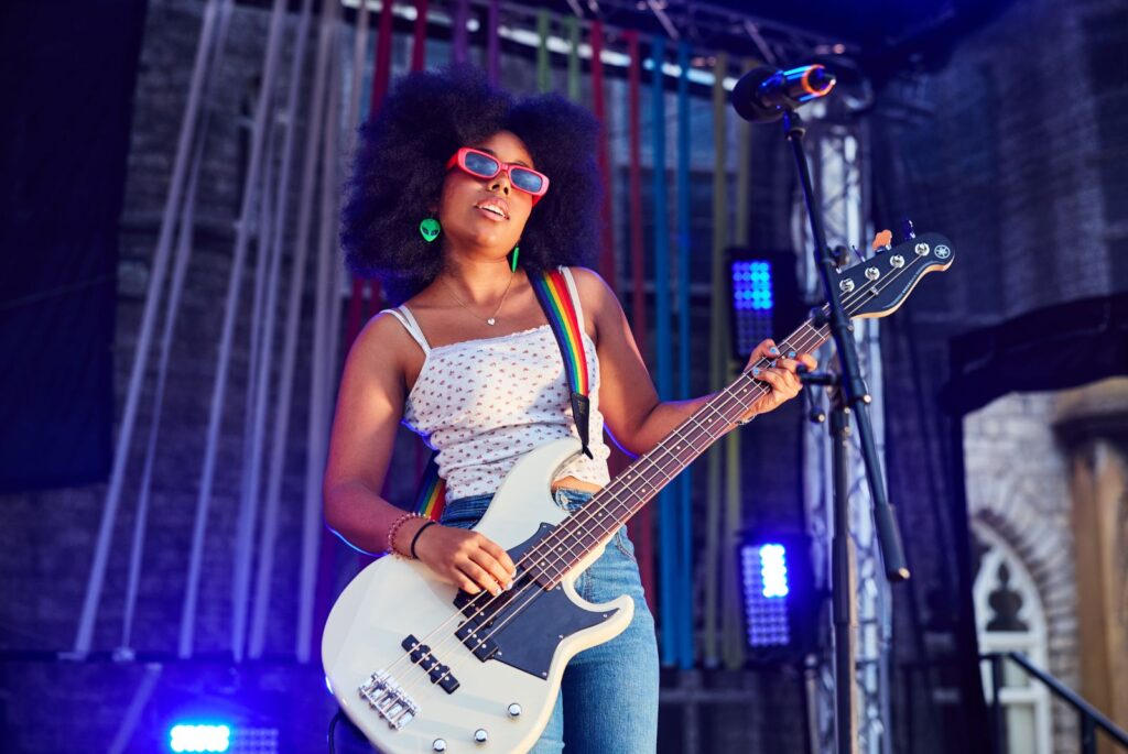 A girl wearing pink sunglasses, a white and pink floral vest, green alien earrings and blue jeans is playing a white guitar with a rainbow strap.