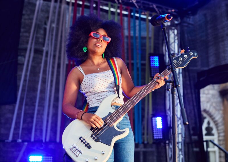 A girl wearing pink sunglasses, a white and pink floral vest, green alien earrings and blue jeans is playing a white guitar with a rainbow strap.
