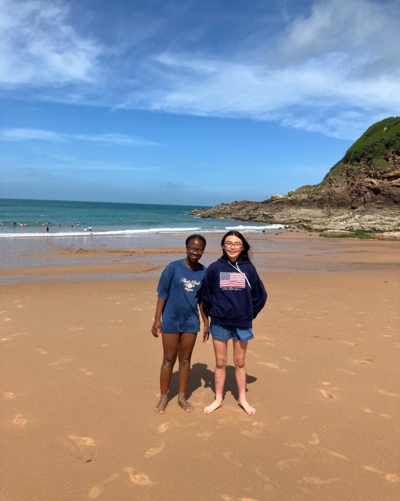 Two UIII pupils stood on a beach in front of the sea. They are both wearing a sweatshirt and denim shorts.
