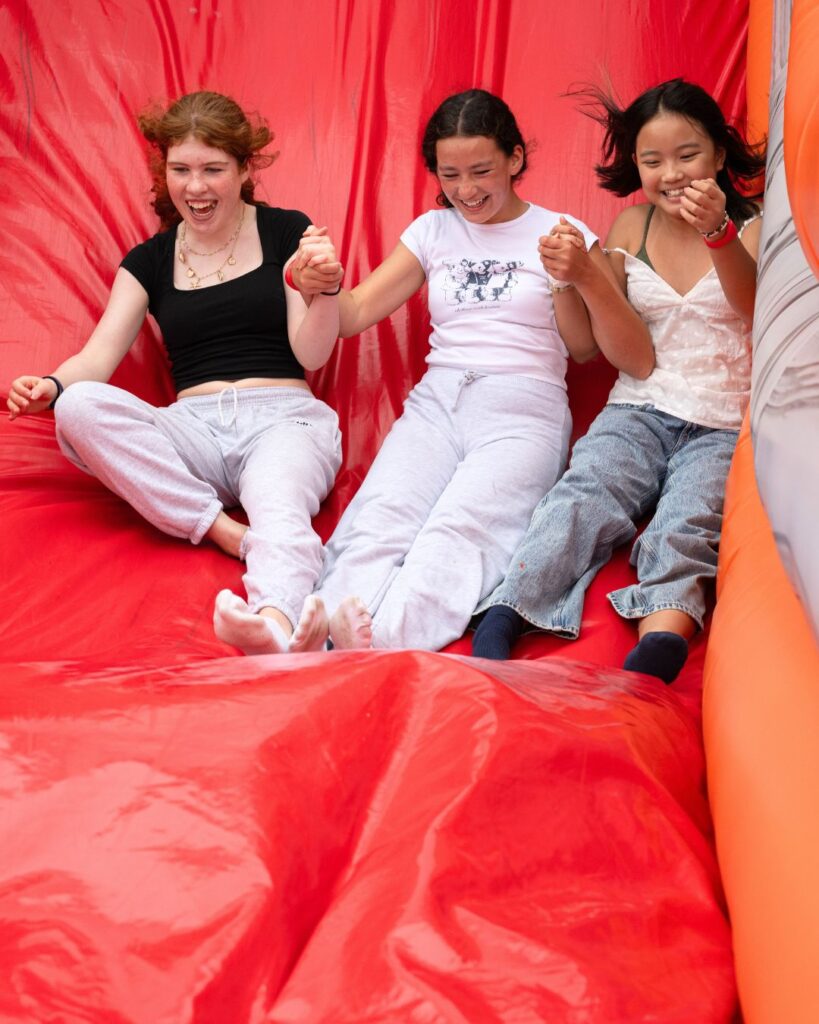 Three girls holding hand, smiling and laughing whilst going down a red inflatable slide.