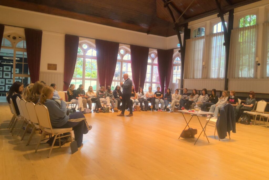 Andrew Wright stood in front of the UV who are sat in a semicircle.
