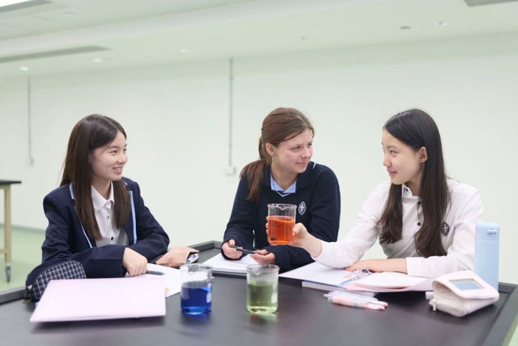 Three pupils sat at a table with a book and a beaker each. One beaker has blue liquid, one has orange, and the other lime green.