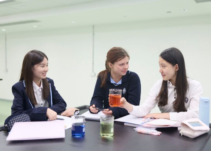 Three pupils sat at a table with a book and a beaker each. One beaker has blue liquid, one has orange, and the other lime green.