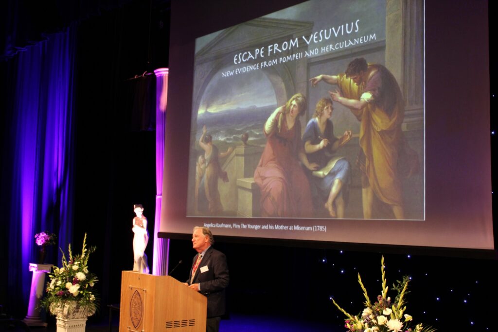 Man standing at plinth in front of a powerpoint presentation on classics