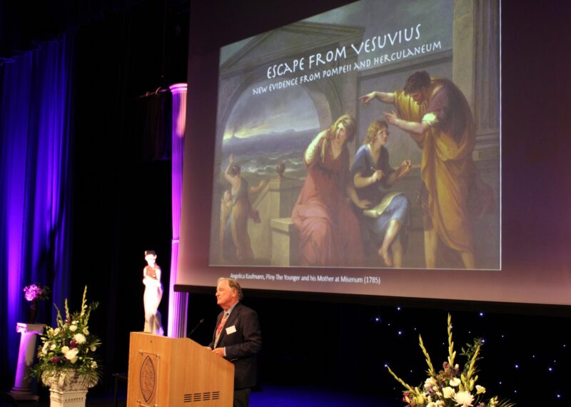 Man standing at plinth in front of a powerpoint presentation on classics