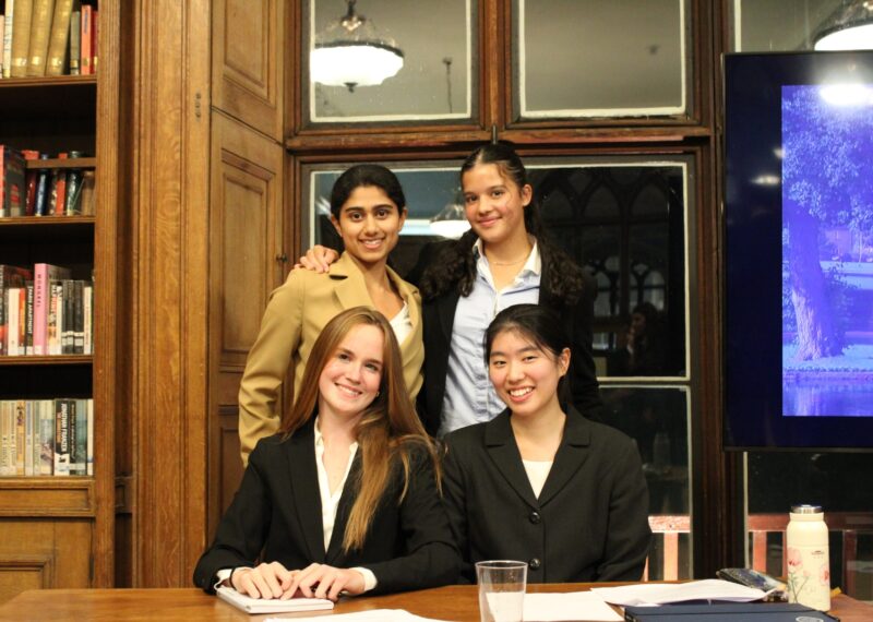 Four Girls, two seated, two standing, at a table in a library posing for the camera wearing suits.